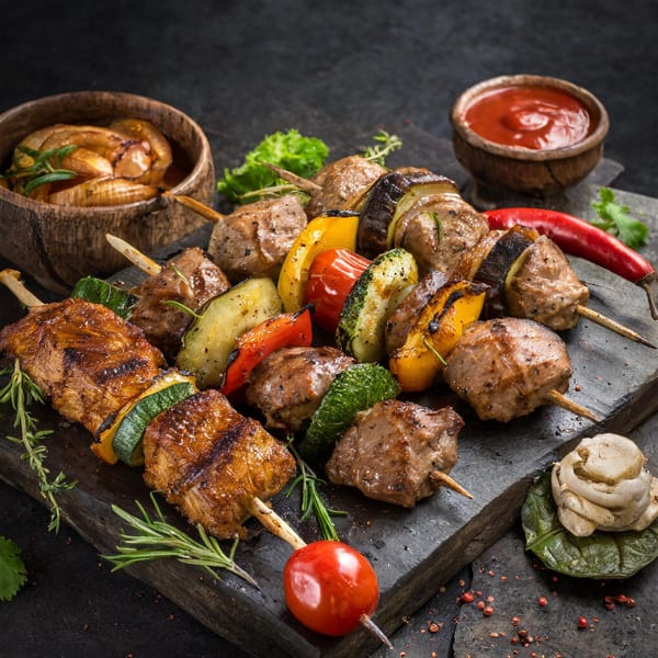 Grilled Middle-Eastern meat and vegetable skewers with sauce and roasted potatoes on a dark board.